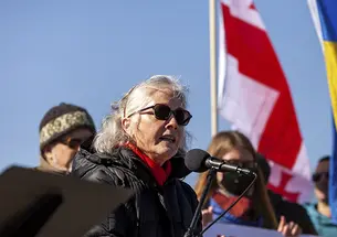 Former U.S. ambassador to Ukraine Marie Yovanovitch ’80 speaks into a microphone during a solidarity rally for Ukraine on Feb. 19, outdoors, wearing sunglasses, with blue sky and the Ukrainian flag in the background.