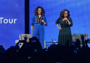 Michelle Obama and Oprah address an audience from a stage.