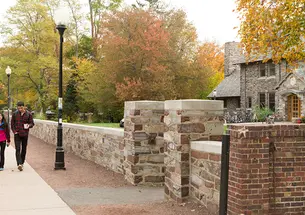 Two students holding coffee cups walk on a sidewalk next to a stone wall. A stone eating club can be seen to the right behind the wall.