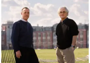 Two men stand on grass with a building far in the background.