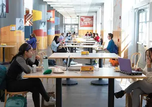 This is a photo of students working on laptops and eating in the Frist Campus Center, distanced by sitting longways across from each other, two to a table.