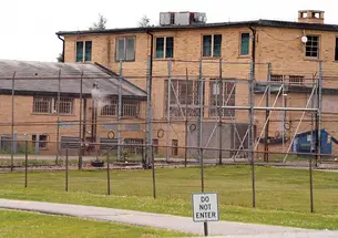 This is a photo of the Edna Mahan Correctional Facility, made of yellow brick with tall chain-link fences in front and a small sign reading "DO NOT ENTER."