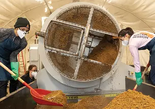 OTC_SCRAP Lab.jpg Students shovel compost that looks like dirt next to a cylindrical machine.