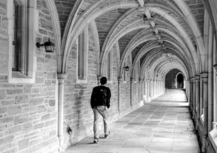 A black-and-white photo of a student walking down an arched stone hallway alone.
