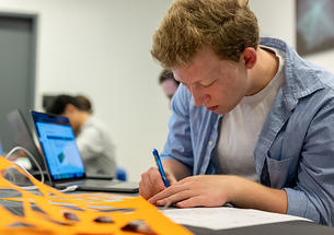A student writes with a pen; a yellow piece of paper with shapes punched out is in the foreground.