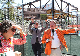 A man in an orange striped blazer speaks into a microphone in front of a construction site.