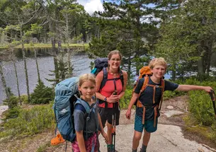 A woman and her two children wear backpacks on a trail; trees and a body of water are in the background.