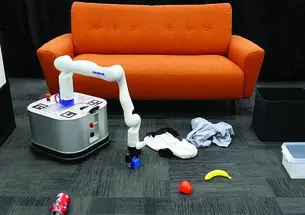 A square robot with a long white arm picks up objects on a carpeted floor in front of an orange couch.