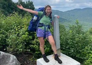 Wearing her backpack and hiking boots, Ruby Platt flings out an arm and smiles as she stands in front of the Green Mountains, holding onto an obelisk-shaped marker that reads "Treaty of Washington."