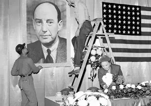 In this black-and-white photo, two men hang a poster of Adlai Stevenson on a wall next to an American flag, while a woman arranges flowers on a long table. Preparations are made for a campaign stop for Adlai Stevenson 1922 in 1952, before he lost to Dwight Eisenhower, 442 electoral votes to 89.
