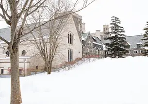 A view of part of Princeton’s Gothic stone campus, viewed up a snowy hill, with two large pine trees at the top.