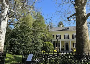 This is a photo of the yellow Maclean house on Princeton's campus, with two large sycamore trees and a wrought iron fence in front.