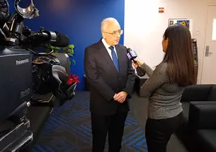 This is a photo of Alain Sanders being interviewed by a news reporter holding a microphone. A video camera filming them is in the foreground..