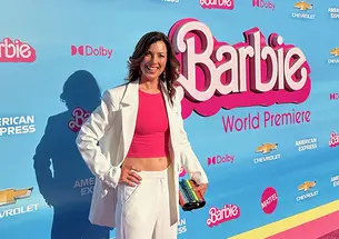 Cate Adams ’08 poses in front of a banner reading "Barbie" at the world premier. She's wearing a pink cropped top under a white suit.