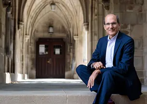 This is a photo of David Nirenberg sitting on the steps of a building with Gothic arches in the background.