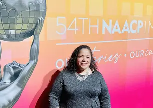 Janice Johnston ’92 stands in front of a large pink-and-yellow sign reading "54th NAACP." Part of the text is cut off. There's also a photo of a silver statue of Atlas holding up the globe.