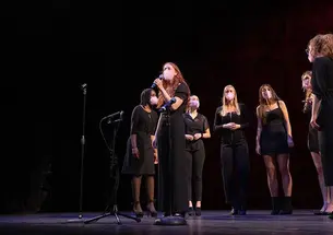 This is a photo of the Tigerlilies singing on stage; eight women are wearing black clothes and pink face masks while they sing.