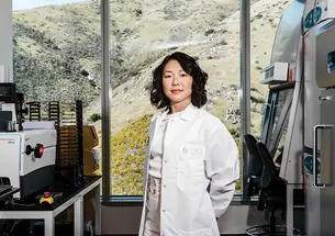 This is a photo of Alice Zhang ’10 inside the Verge Genomics laboratory in South San Francisco, wearing a lab coat and surrounded by machines, with a view of scrubby vegetation through the window behind her.