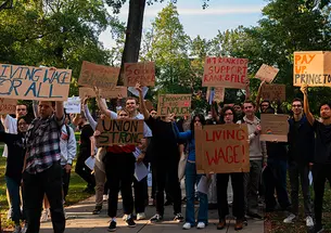 A crowd standing outside holds signs reading "Living Wage!" and "Pay Up Princeton" and #1 Ranked? Support Rank & File" and "Union Strong."