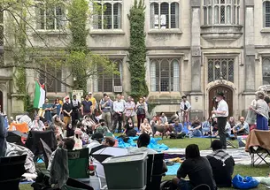 A man stands at a microphone surrounded by protesters; a Gothic building is in the background.