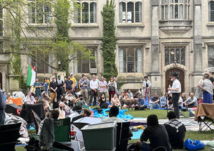 A man stands at a microphone surrounded by protesters; a Gothic building is in the background.