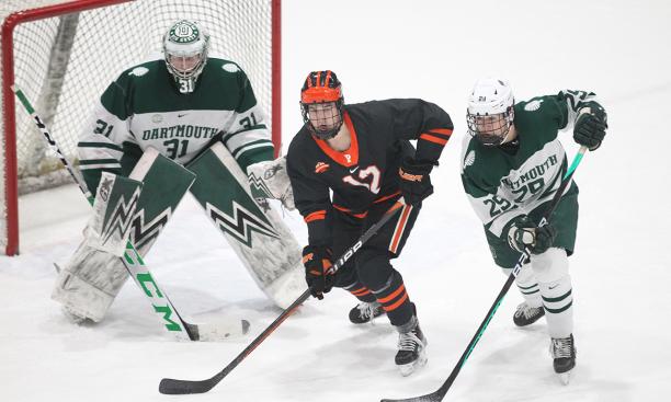 A Princeton men’s ice hockey player skates between a Dartmouth player and the Dartmouth goalie, with the goal in the background. 