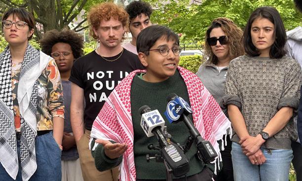 Wearing a red and white keffiyeh around her shoulders, Aditit Rao speaks into two microphones labeled "news 12" and "6 abc." Six other students stand behind her.