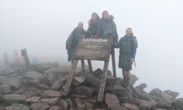 Four people in rain jackets cluster in thick fog around a wooden sign reading “Katahdin,” perched on a pile of rocks.