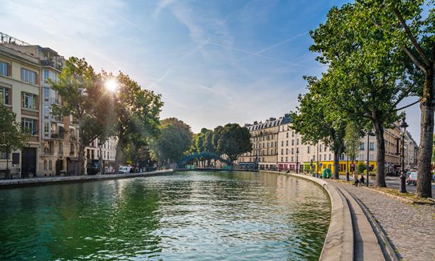 A canal curves around a Paris street with the sun visible through trees in the background.