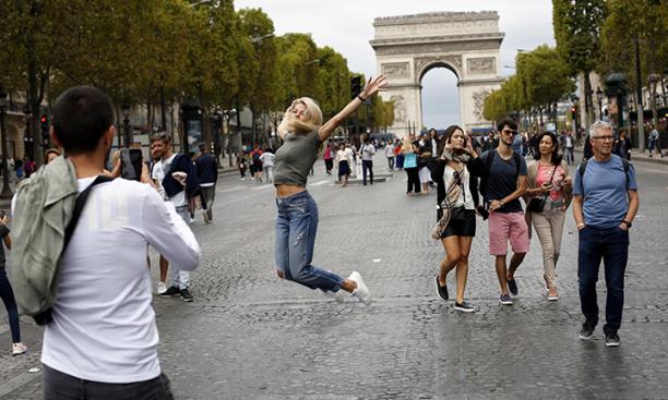 A woman jumps in the air on the street in front of a large stone arch monument in Paris.