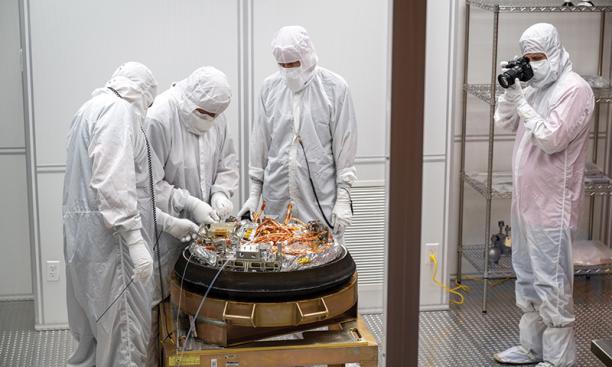 Four people in white hazmat suits stand around the sample from asteroid Bennu as it's unpacked 
