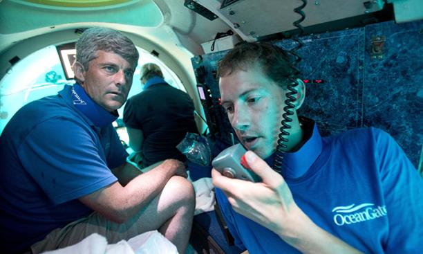 Three men in a small submersible; the one in the front is speaking into a radio handset and wearing a blue shirt with the OceanGate logo.