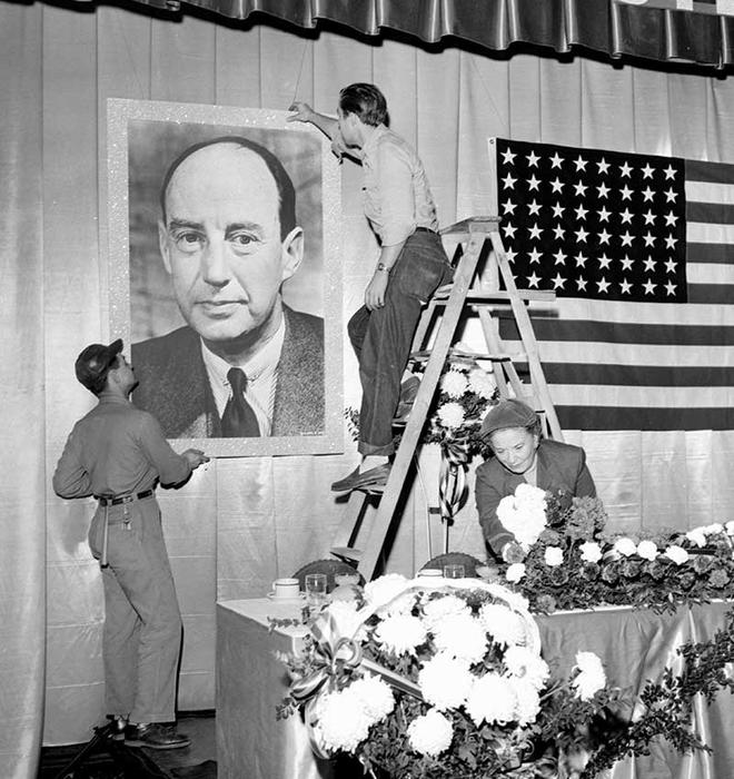 In this black-and-white photo, two men hang a poster of Adlai Stevenson on a wall next to an American flag, while a woman arranges flowers on a long table. Preparations are made for a campaign stop for Adlai Stevenson 1922 in 1952, before he lost to Dwight Eisenhower, 442 electoral votes to 89.