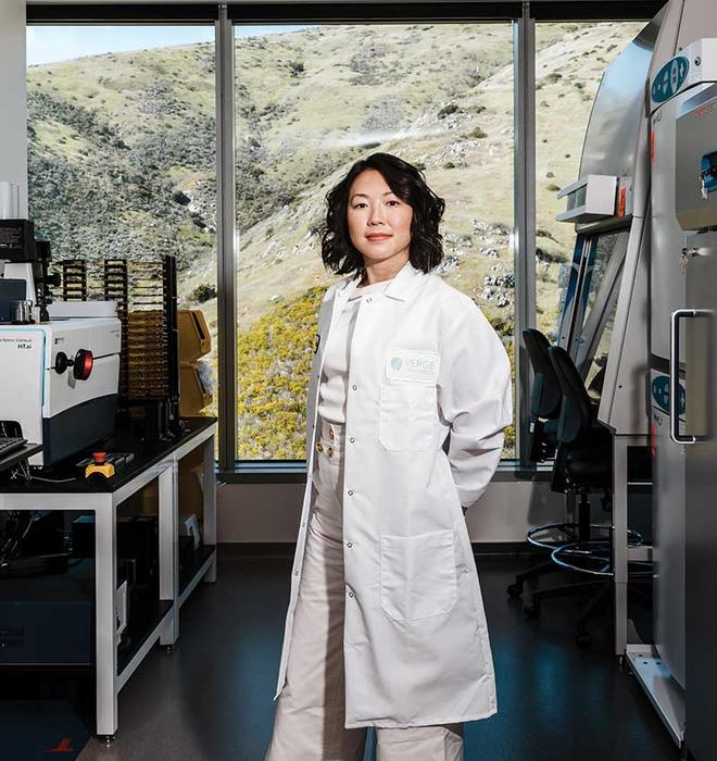 This is a photo of Alice Zhang ’10 inside the Verge Genomics laboratory in South San Francisco, wearing a lab coat and surrounded by machines, with a view of scrubby vegetation through the window behind her.