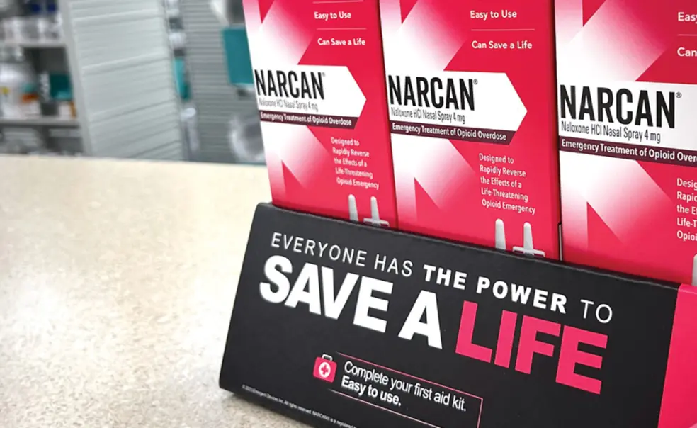 OTC_Narcan Student Overdoses Public Health.jpg Pink boxes of Narcan tacked on counter with a sign that reads Save a Life