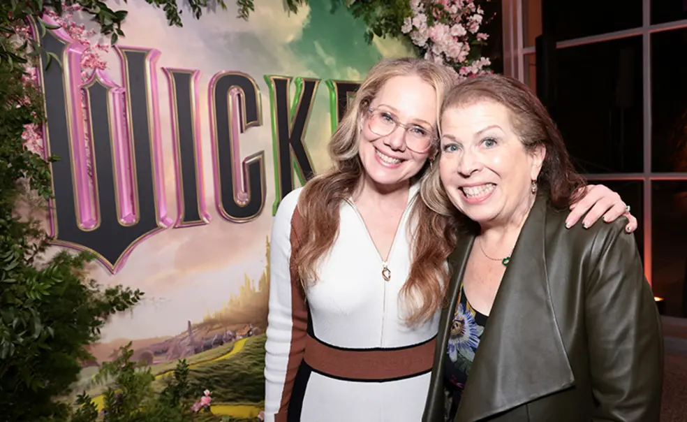 TOW_WinnieHolzman.jpg Two women smile for the camera in front of a poster for "Wicked" the movie.