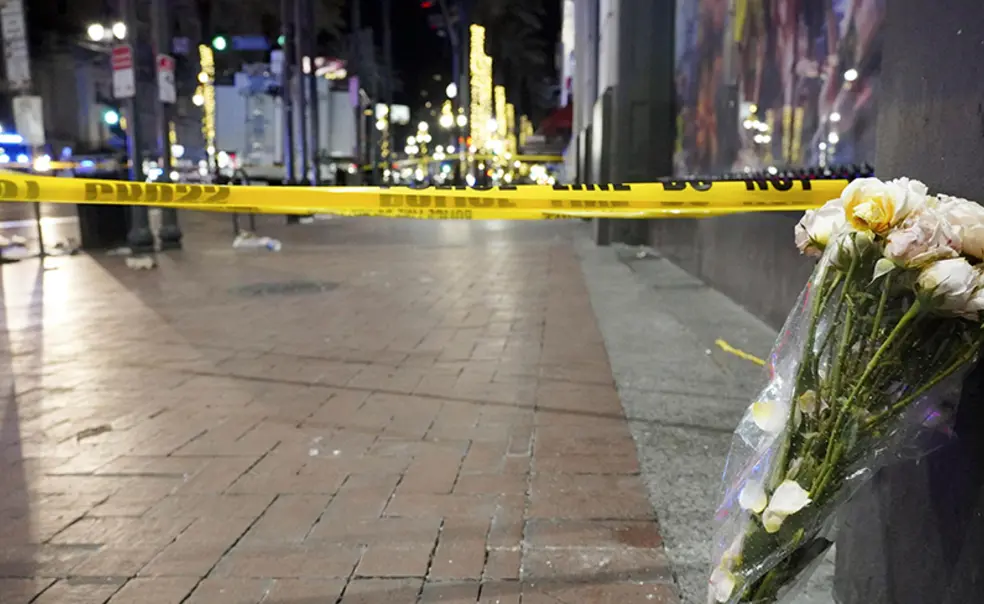 TigerBech_new orleans truck attack.jpg At night, a bouquet of flowers leans up against a building next to a brick sidewalk blocked off with caution tape.