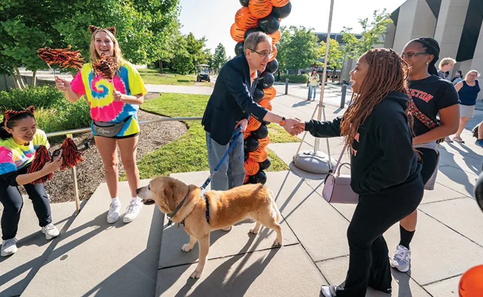 President Eisgruber shakes a woman's hand in front of an orange-and-black balloon arch; his other hand holds the leash of a yellow labrador.