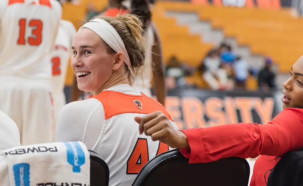 WholeStudent_Reyneke_game.jpg A Princeton basketball player smiles as she looks behind her from the bench on the side of a game.
