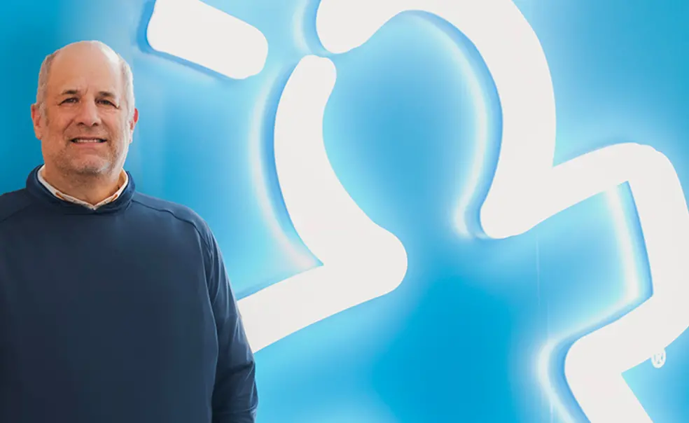 Rick Woldenberg at Educational Insights Headquaters.jpg Rick Woldenberg ’81 against a blue background where his company's logo — a person with lines like a lighbulb overhead — is glowing in white neon bulbs.