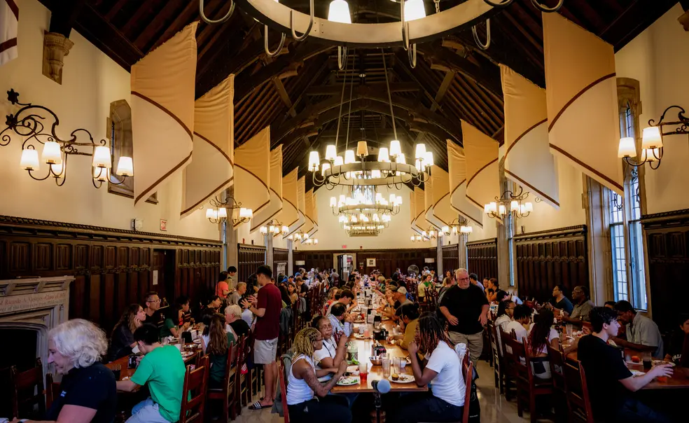 Students sit at long tables arranged in three rows in a dining hall with a high wood ceiling.