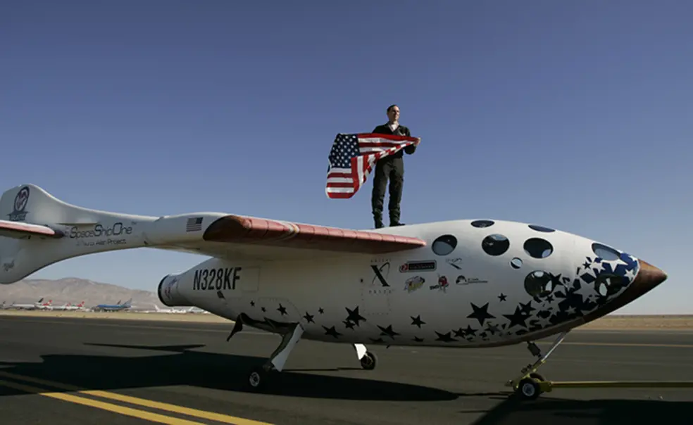AITN_BrianBinnie.jpg Brian Binnie *78 stands on what looks like a small airplane, holding an American flag.