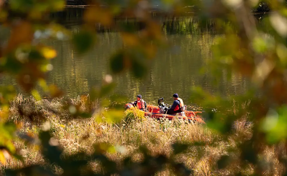 Boat.jpg Seen through an opening in the trees, two people in orange vests motor on Lake Carnegie in an orange boat.
