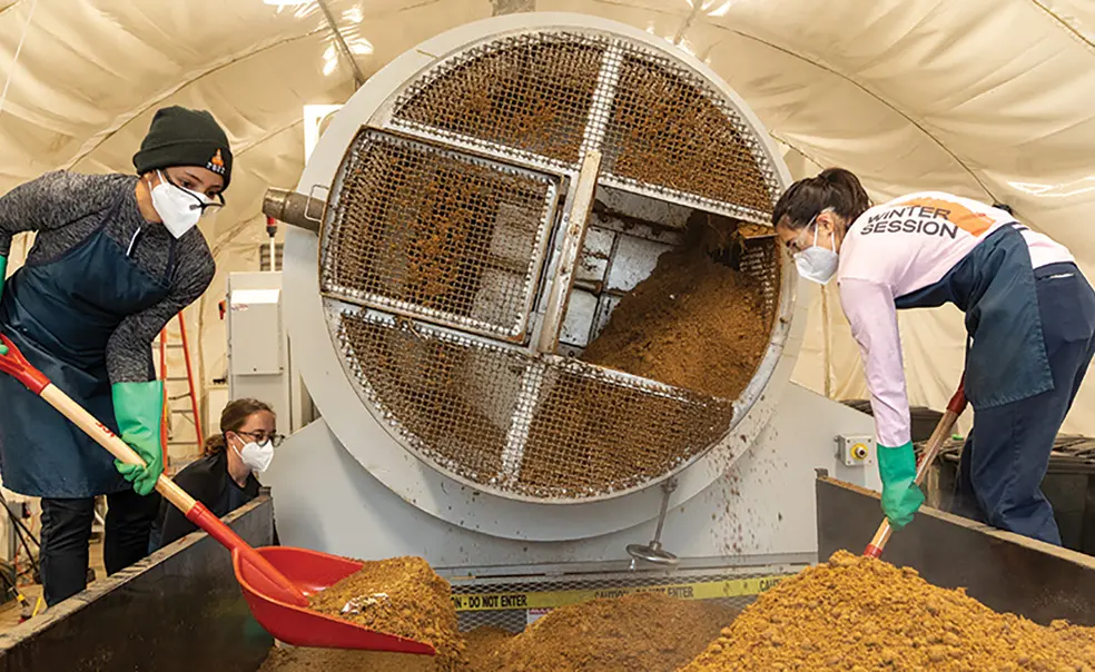 OTC_SCRAP Lab.jpg Students shovel compost that looks like dirt next to a cylindrical machine.