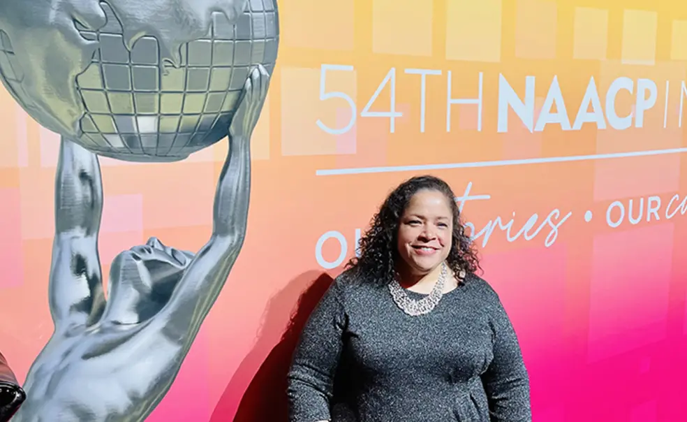 TOW_JaniceJohnston.jpg Janice Johnston ’92 stands in front of a large pink-and-yellow sign reading "54th NAACP." Part of the text is cut off. There's also a photo of a silver statue of Atlas holding up the globe.