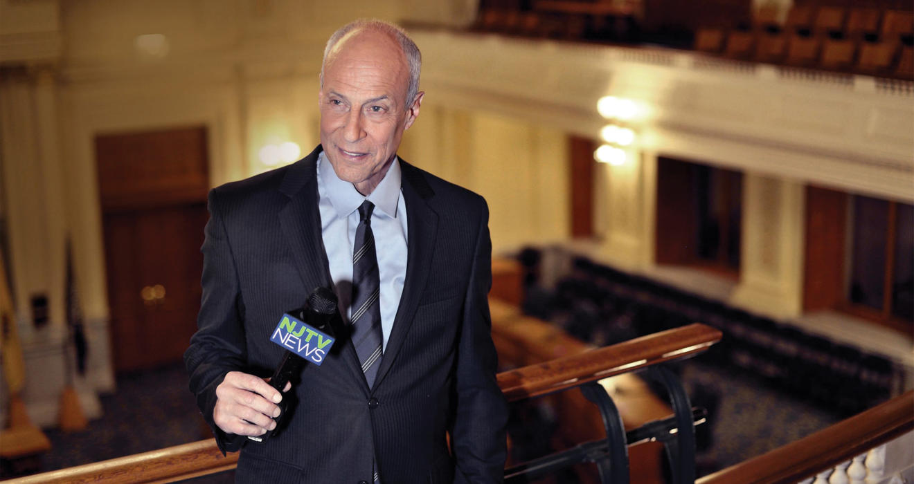 Michael Aron *70 holds a microphone while speaking from an upper level of the NJ statehouse.