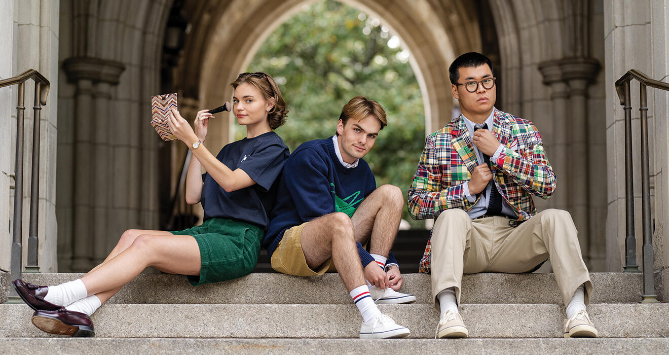 Three students sit on steps outside, with a pointed arch in the background.