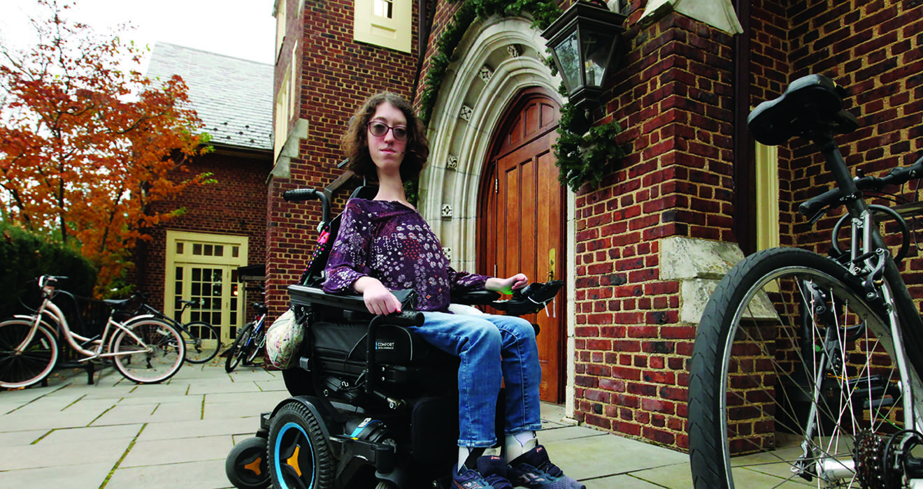 This is a photo of Naomi Hess ’22 in her motorized wheelchair in front of a Princeton campus building. Bicycles are in the foreground and background.