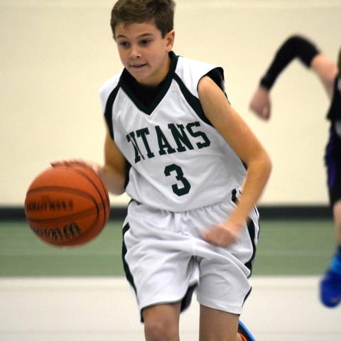 Young Caden Pierce, in a white jersey with green text on it that reads Titans and is labeled with a 3, dribbles the ball with a defender in pursuit. 