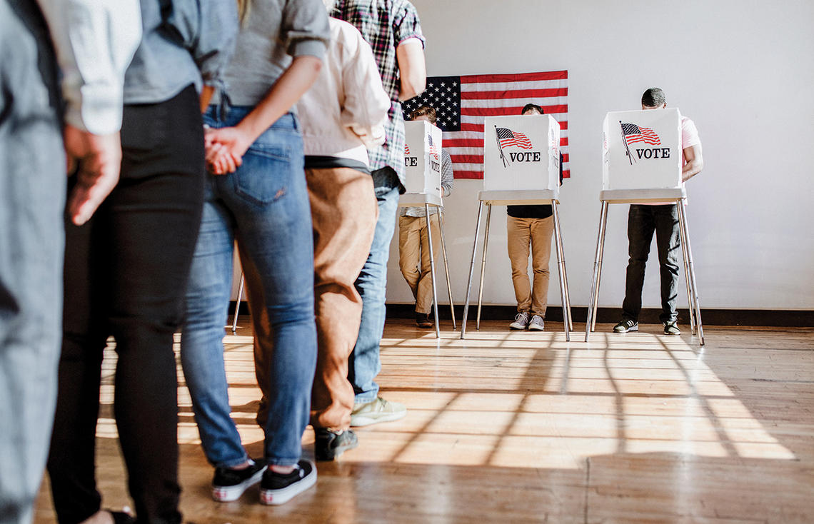 A line of people waiting to vote is shown from behind, from the waist down. People vote in the background.